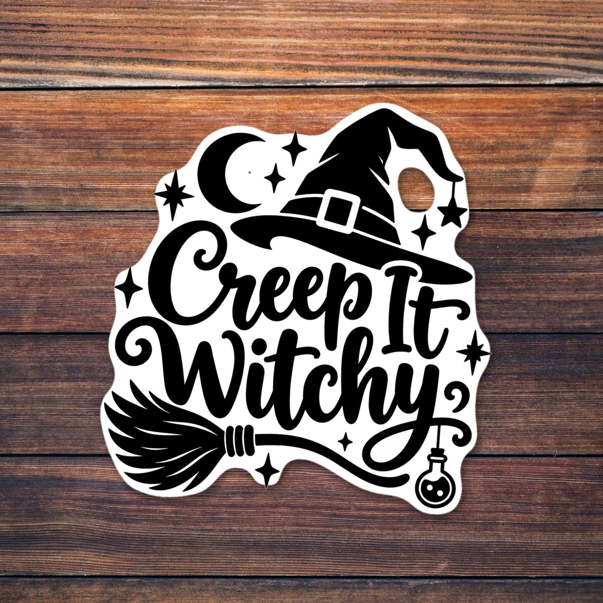 Black script text reading Creep It Witchy with a buckled witch hat on top, a crescent moon, sparkle stars, a hanging star, a broomstick underneath and a small hanging potion bottle shown on a wooden table