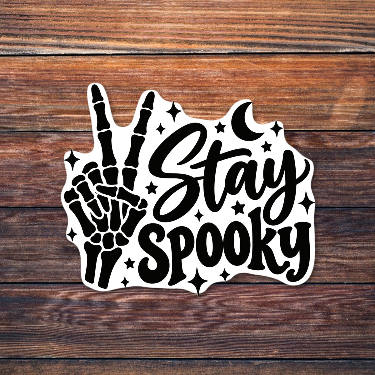 Black design of a skeleton hand making a peace sign next to the script words Stay Spooky, with a crescent moon, stars and sparkles around it shown on a wooden table