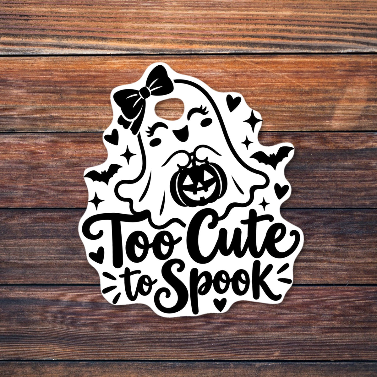 Black and white line art of a smiling cartoon ghost with a large hair bow and eyelashes holding a jack-o'-lantern trick-or-treat bucket, surrounded by bats, hearts, stars and sparkles, with the script text "Too Cute to Spook". shown on a wooden table